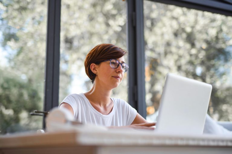 A woman wearing glasses looks at her laptop near a bright window, engaging in an online mental health class.
