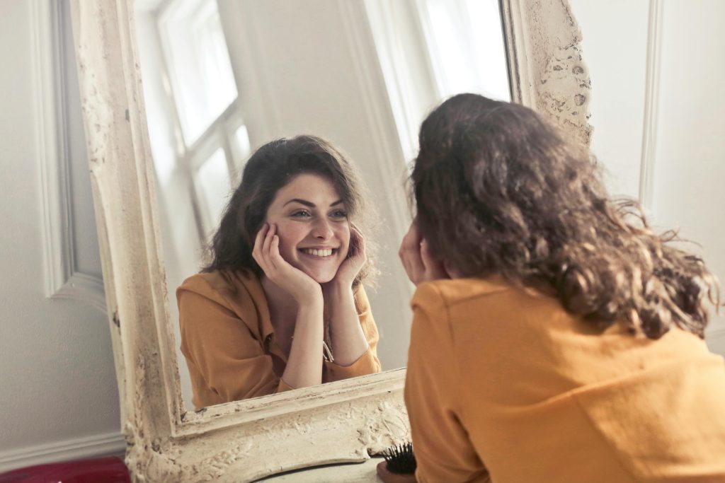 Photograph of a smiling young woman with wavy brown hair wearing a mustard yellow top, looking at her reflection in a large, vintage-style mirror.