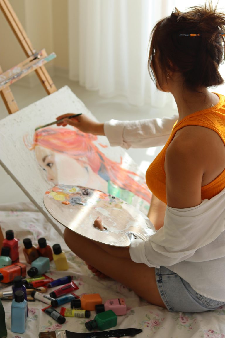 A woman sitting on the floor working on a colorful portrait painting, showcasing her passionate engagement with fine art.