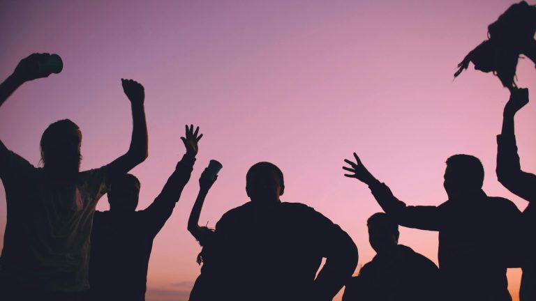 Silhouettes of a group of mental health friends celebrating and raising their drinks against a vibrant purple and pink sunset sky.