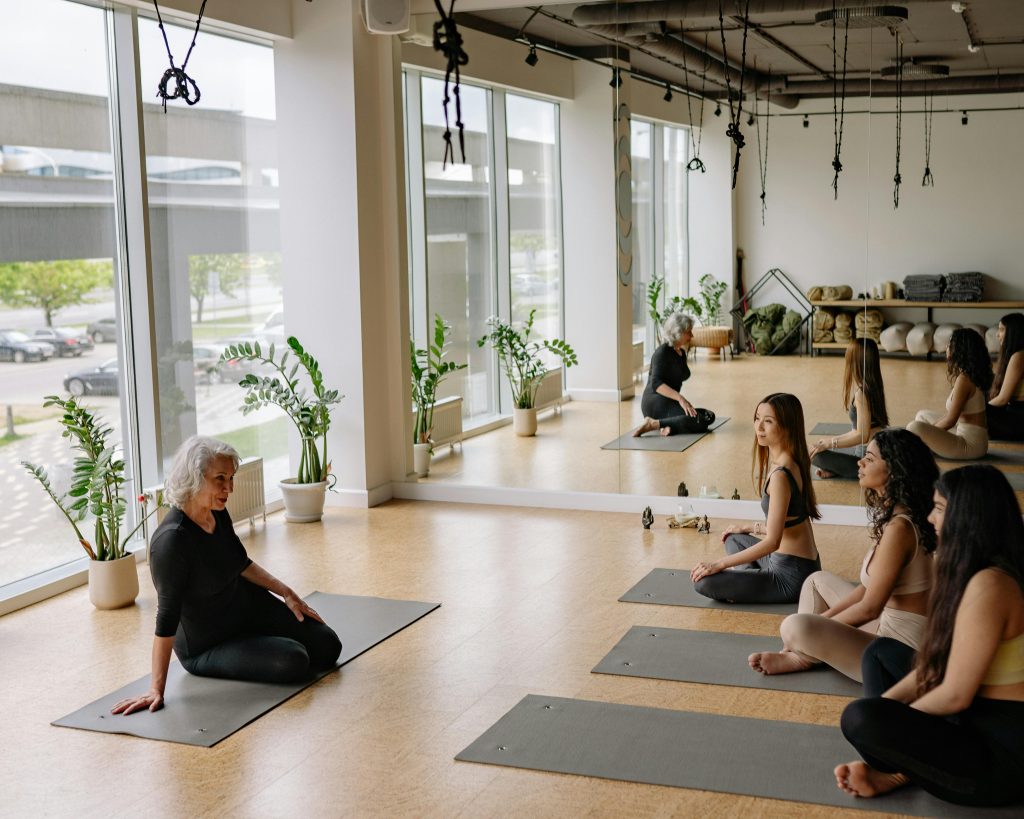 An elderly instructor leading a yoga group class in a bright studio with large windows.