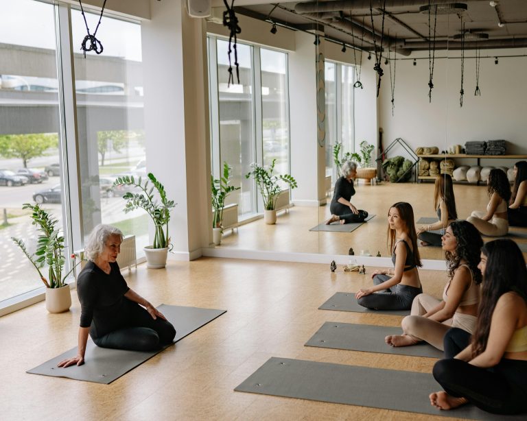 An elderly instructor leading a yoga group class in a bright studio with large windows.