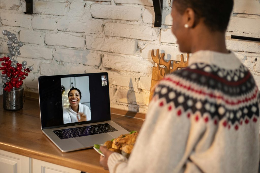 A woman in a holiday sweater on a video call via laptop in a kitchen, holding a plate of food during a festive celebration.