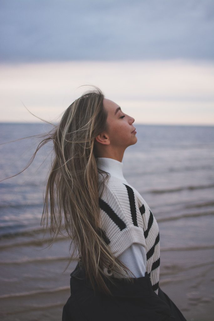 Photograph of a woman from the side with her eyes closed, standing on a beach with long hair blowing in the wind.