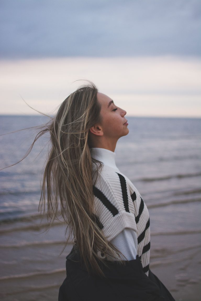 Photograph of a woman from the side with her eyes closed, standing on a beach with long hair blowing in the wind.