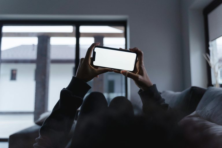 Photograph of a person lying down in a dimly lit room, holding a smartphone horizontally with a bright, glowing white screen.