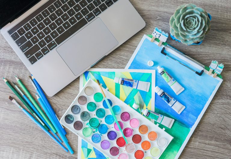 Artist workspace with watercolors and laptop, showcasing types of art medium on a wooden desk.