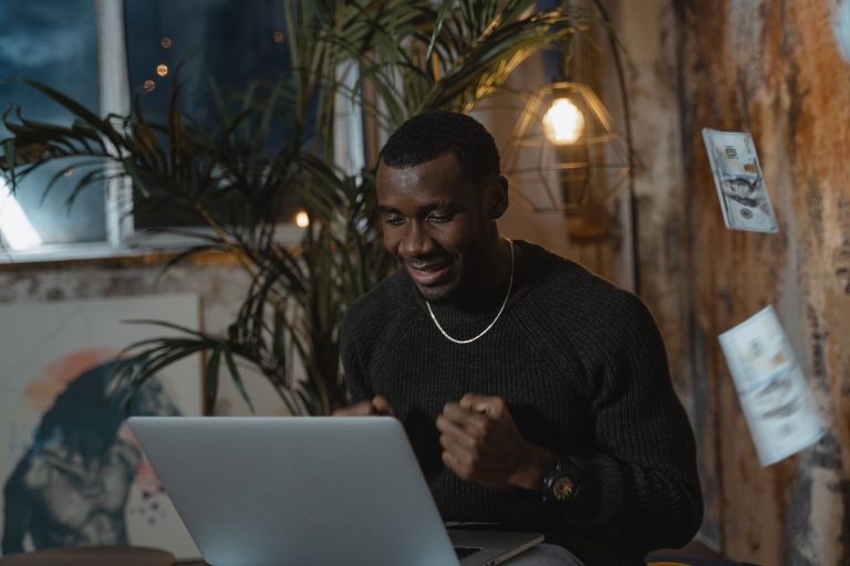 Smiling online teacherpreneur celebrating in front of his laptop while US dollar bills fall around him.