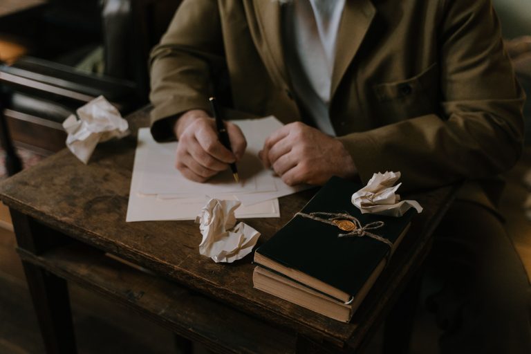 Person writing on paper with a fountain pen at a wooden desk with crumpled papers and journals, illustrating how to write a poem.