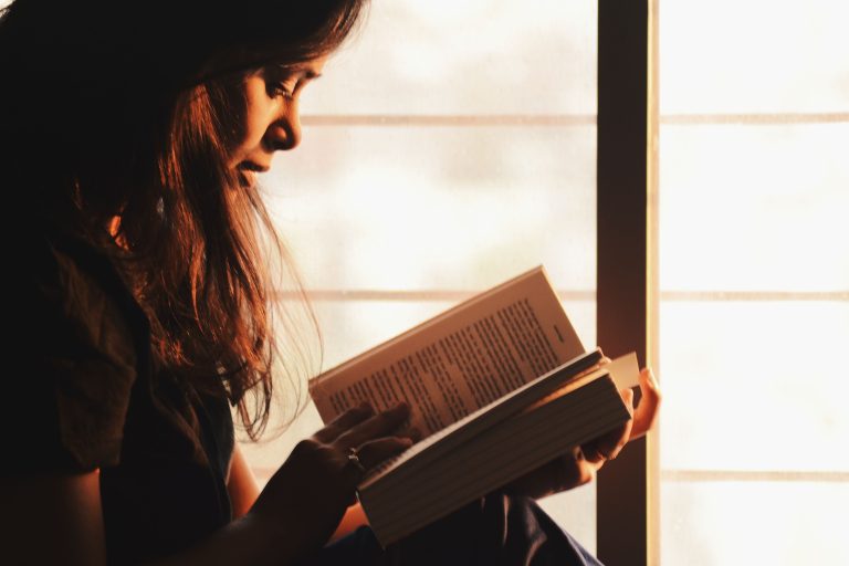 Woman reading a book beside a bright window illuminated by warm sunlight.