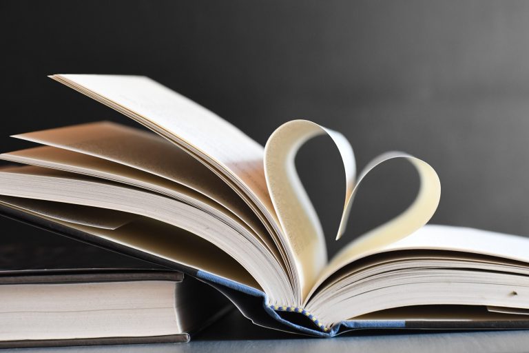 An open book with pages folded into a heart shape, resting on top of another closed book against a dark background.